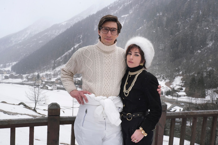 Lady Gaga and Adam Driver in snow. She wears a fur hat, black turtleneck jumper and gold chains, he a cream jumper and ski pants