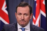 A close up of Mark McGowan in focus with a blue curtain and flags blurred in the background.