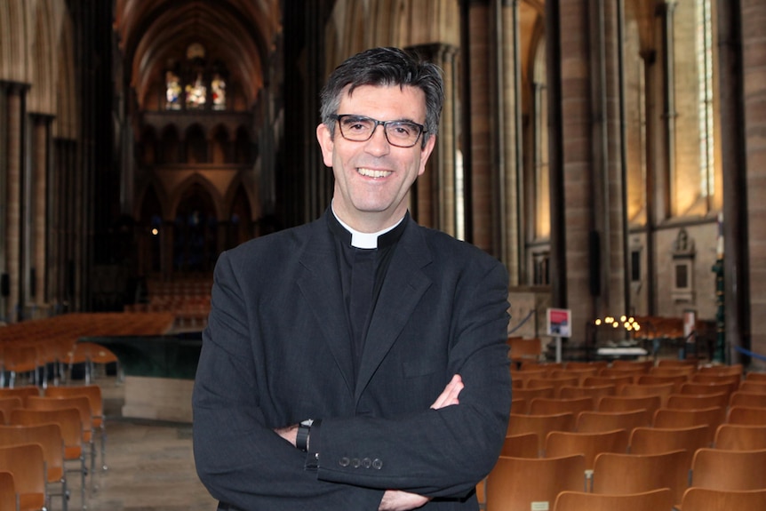 A man with dark hair and glasses, in clerical collar, poses in a church setting