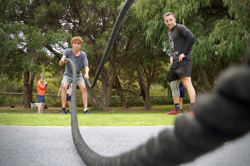 A young man uses the fitness tool battle ropes while a trainer guides him. They are outside in a park and wear fitness attire.