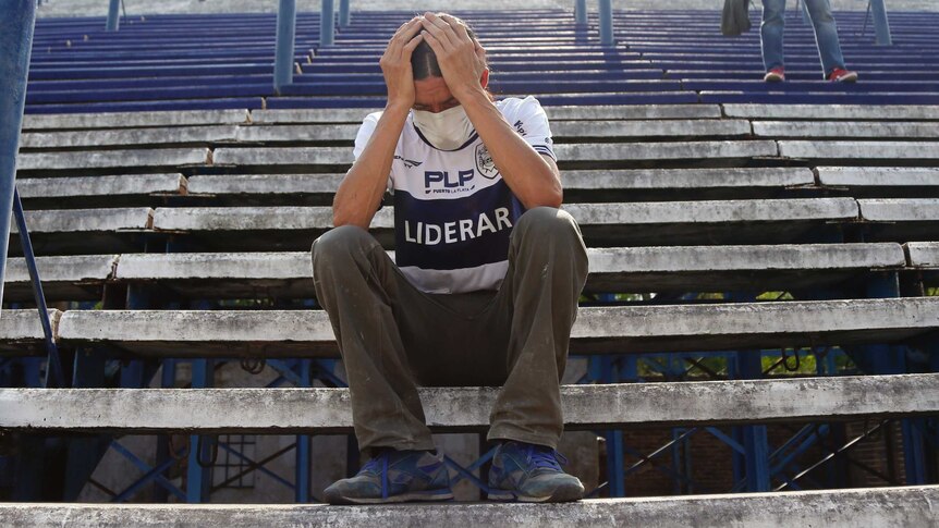 A man holds his head in his hands while sitting in empty bleachers. He is wearing a football jersey and is crying.