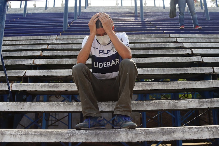 A man holds his head in his hands while sitting in empty bleachers. He is wearing a football jersey and is crying.