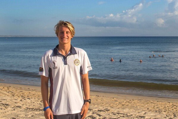 Blonde-haired boy in white t-shirt standing on beach smiling.
