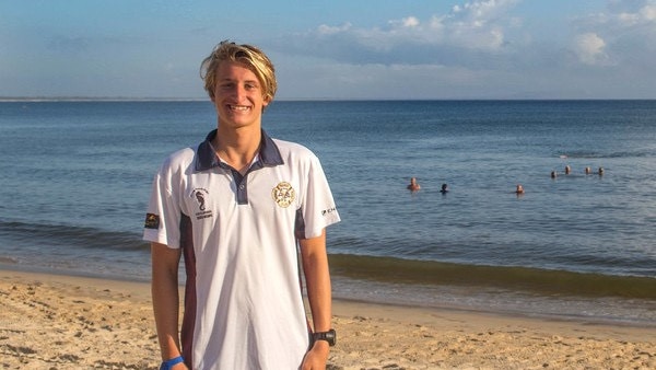Blonde-haired boy in white t-shirt standing on beach smiling.
