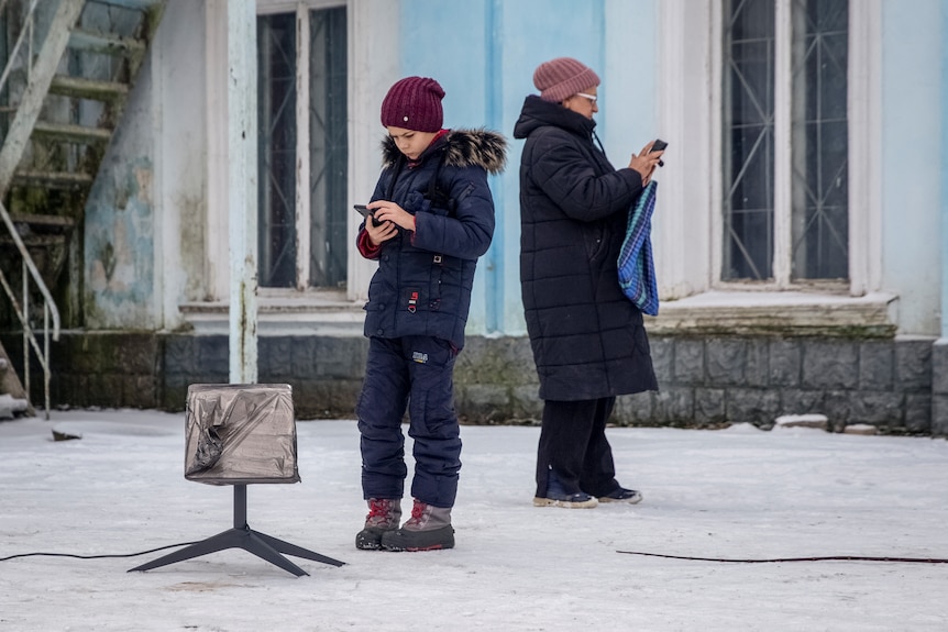 Two people stand in the snow staring at their phones.