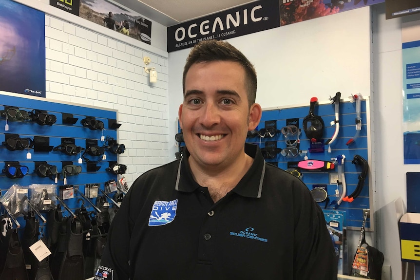 Man smiles in front of scuba diving gear for purchase that hangs on the wall
