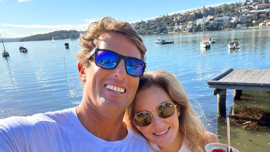 Anthony Carroll takes a selfie by the water with his wife on a sunny day, boats and a wharf in the background.