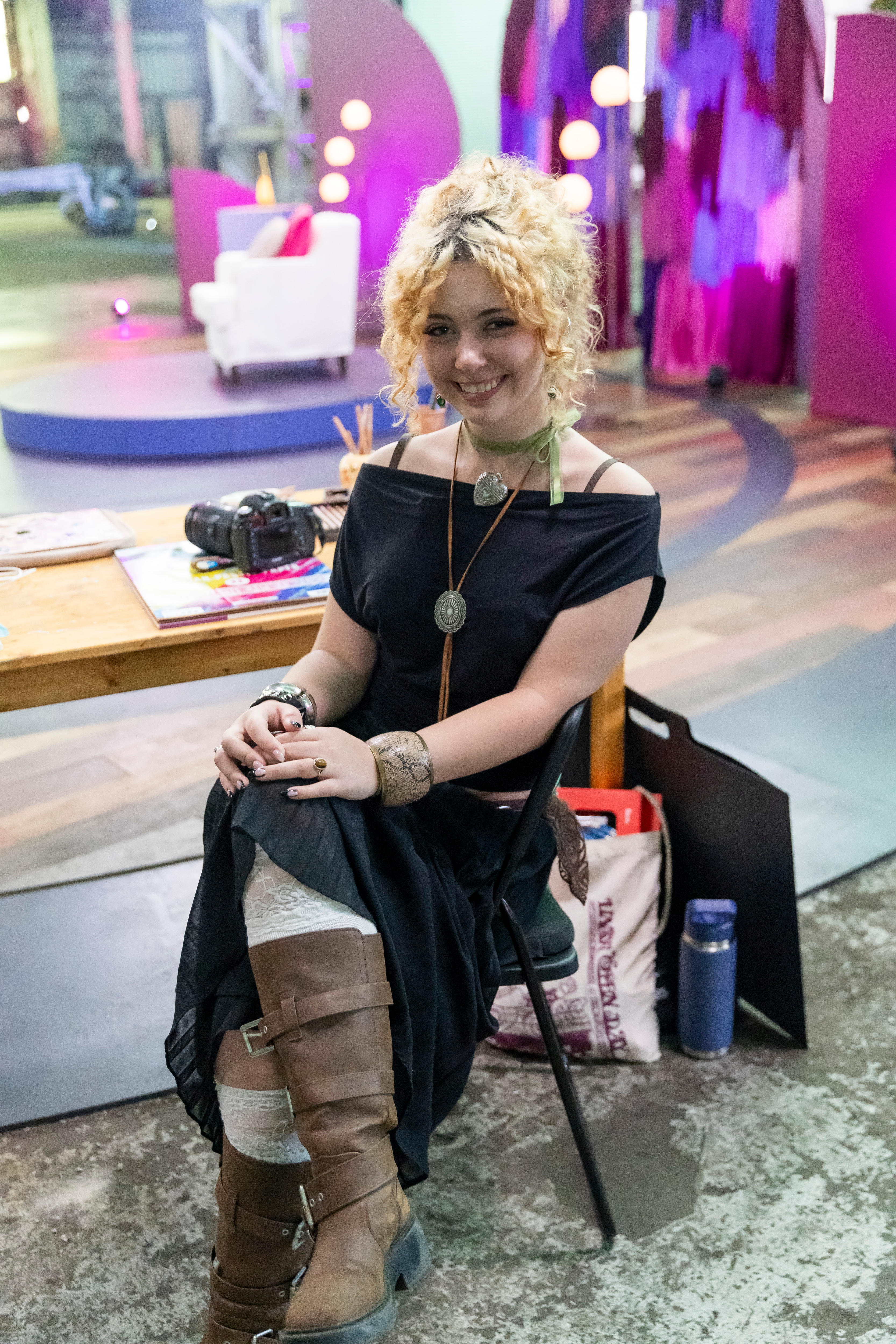 A young woman with curly blonde hair tied back smiles at the camera. A desk with a camera is behind her