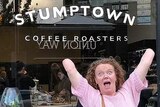 A woman poses in front of a shop called 'Stumptown coffee', gleefully raising her arms, which are both amputated at the elbow.