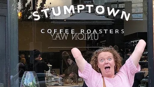 A woman poses in front of a shop called 'Stumptown coffee', gleefully raising her arms, which are both amputated at the elbow.