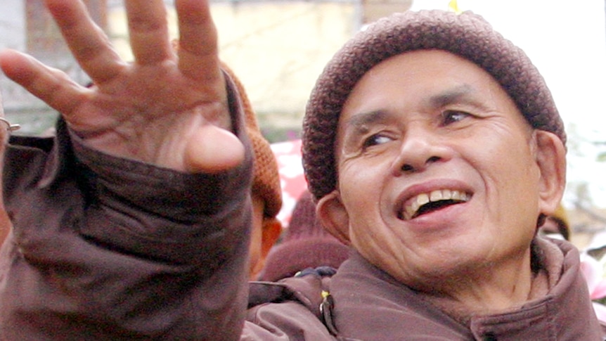 A monk wearing a brown jacket and hat smiles and waves