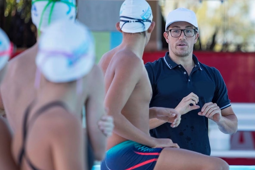 Man in white cap and glasses, speaking to swimmers preparing to dive into pool