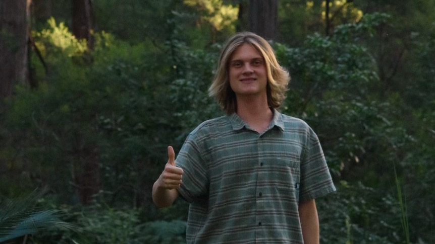 A boy with long dark blond hair standing in a forest smiling, with his thumbs up, wears a check blue shirt.