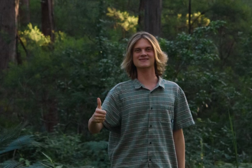 A boy with long dark blond hair standing in a forest smiling, with his thumbs up, wears a check blue shirt.