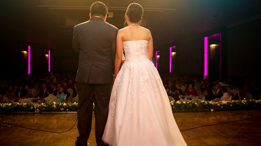 Married couple stand on stage in front of the crowd at their reception.