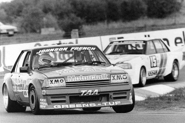 A black and white photo of two muscle cars tearing around a racetrack.