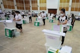 Volunteers check equipment and prepare ballot boxes spaced out in a large hall.
