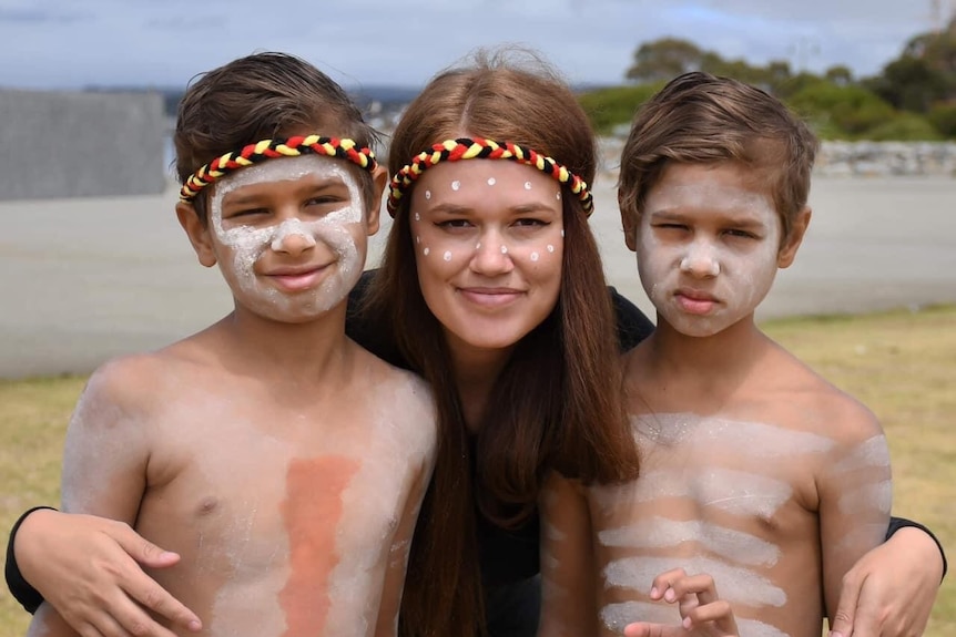 A young woman with her arms around two young boys, all three of them have their faces painted in Aboriginal design.