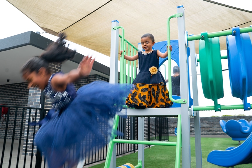 A young girl jumps while a toddler stands behind her on play equipment.
