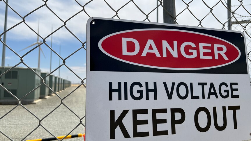 Sign on a chicken-wire fence saying danger, with big battery and wind turbine in the background