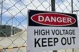 Sign on a chicken-wire fence saying danger, with big battery and wind turbine in the background