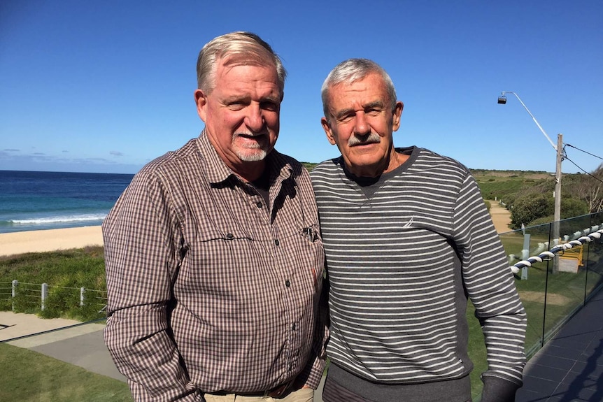 Two men stand on a balcony with the beach behind them