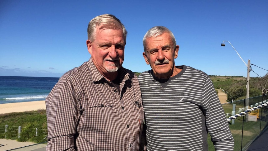 Two men stand on a balcony with the beach behind them