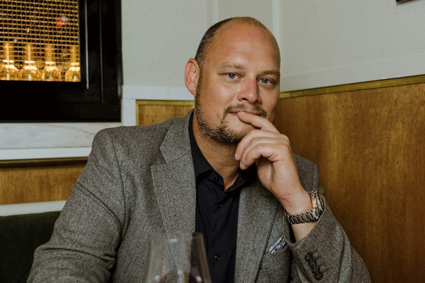 A man with a close shaved head sitting at a table holding a wine glass with his fingers to his mouth.