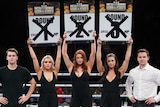 Three ring girls hold up round number signs with crosses through them, standing between two male