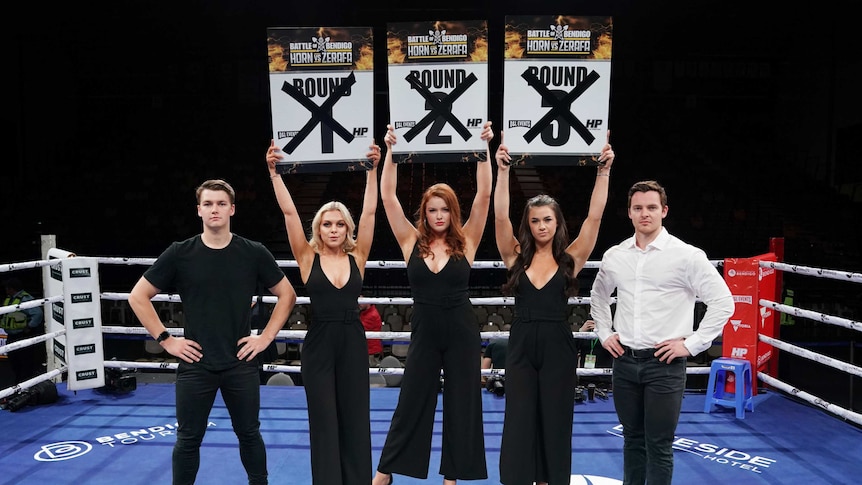 Three ring girls hold up round number signs with crosses through them, standing between two male