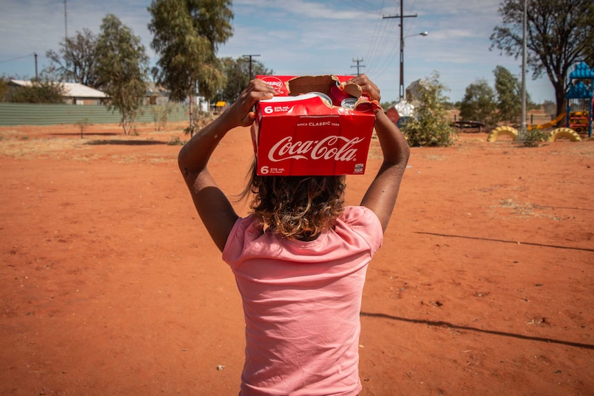 Young girl carries a carton of soft drink through a desert community.