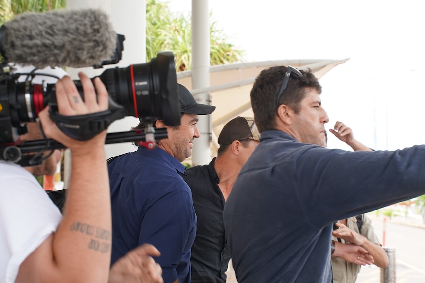 A group of men walk in a close group. A person holding a news camera in filming them. 