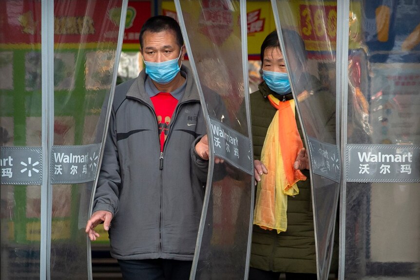 two people in blue face masks walk through a plastic curtain at a grocery store
