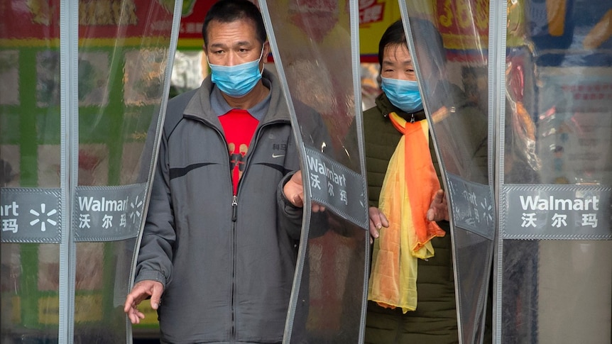 two people in blue face masks walk through plastic curtain at a grocery store