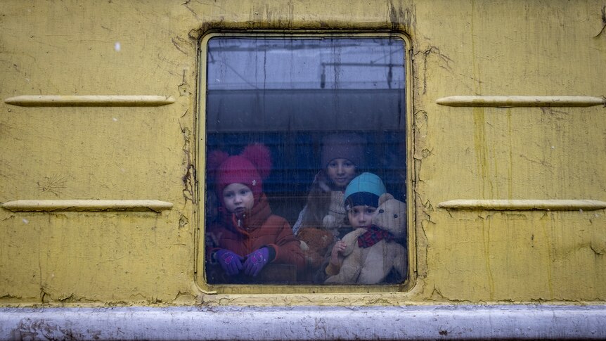 Three children snuggle together in the window of a yellow train. They are wearing winter coats and beanies, one holds a teddy