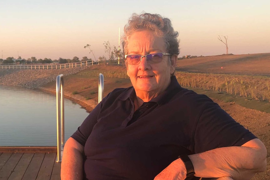 An aged lady with glasses and short curly hair sits on a park bench, behind her is a lake.