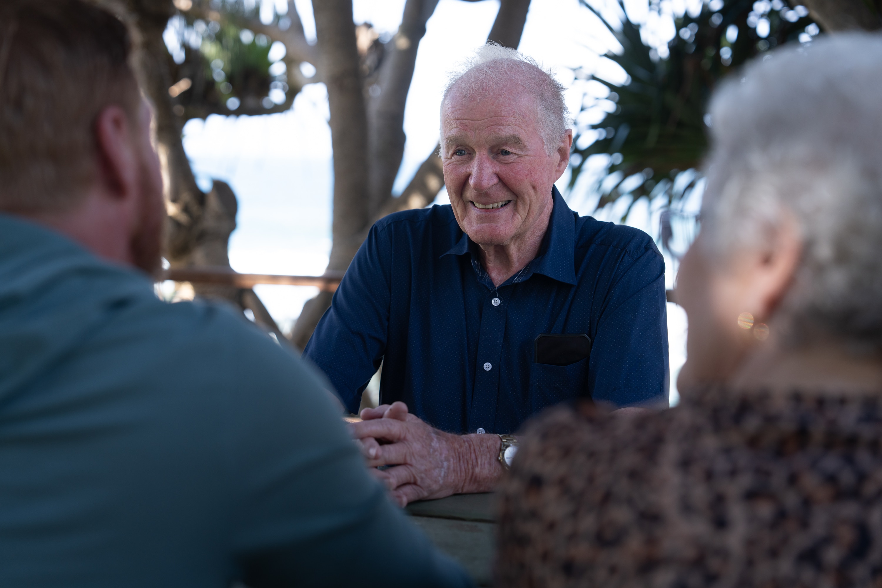 A man smiles sitting at a table outside opposite two people.