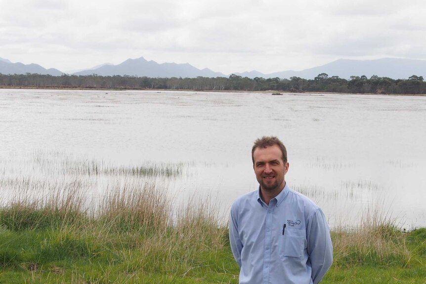 A man in a chambray shirt stands in front of grass and a large body of water.