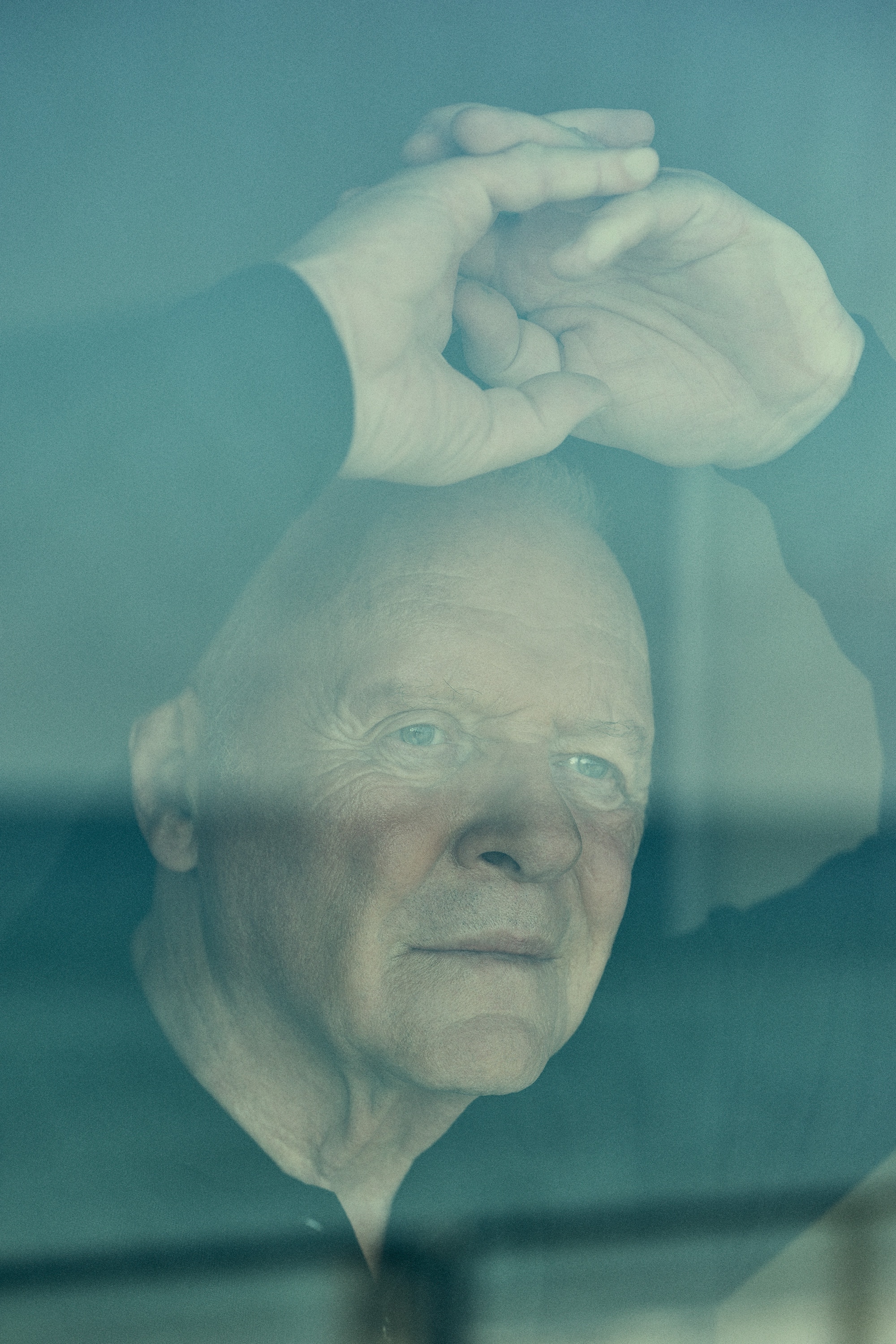 Anthony Hopkins seen through a window, hands raised above head, with soft reflections and muted blue tones.