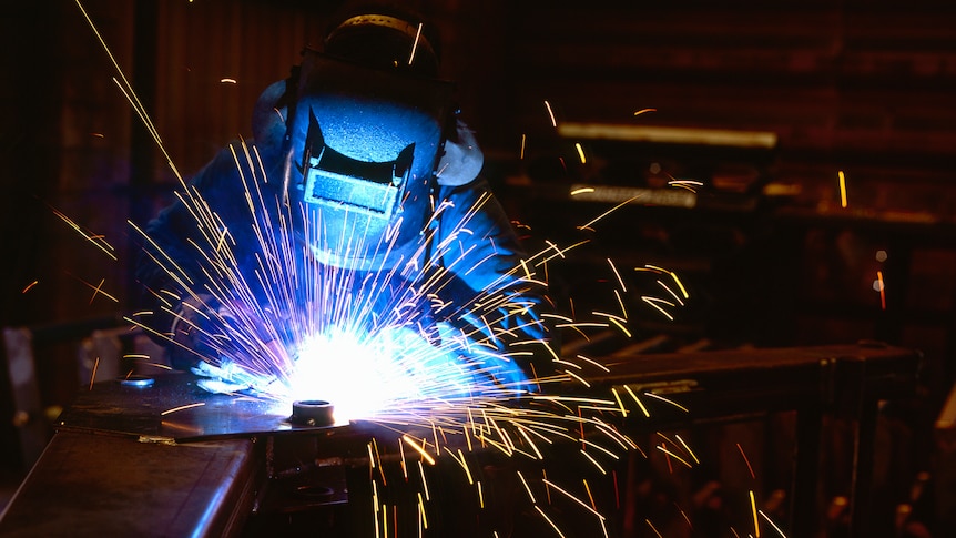 A person in black protective gear welds, as sparks fly.