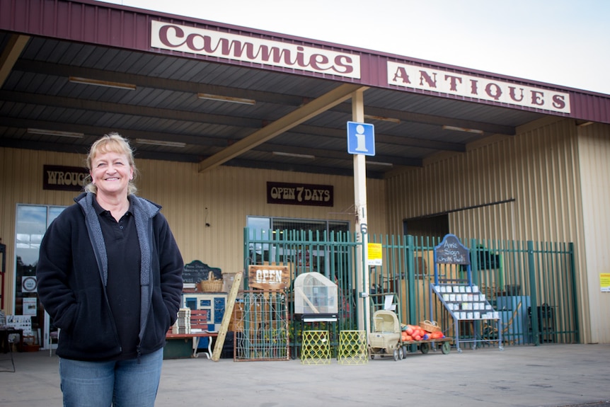Cathie O'Toole stands in front of a shed displaying the sign Cammies Antiques and filled with furniture and collectables.