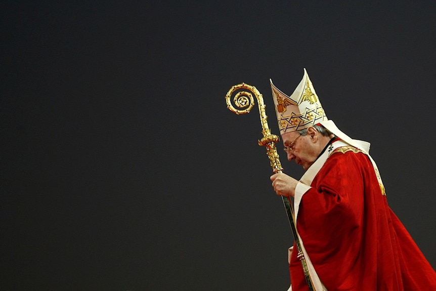 Pell, wearing red religious robes, looks down as he carries an ornate staff.