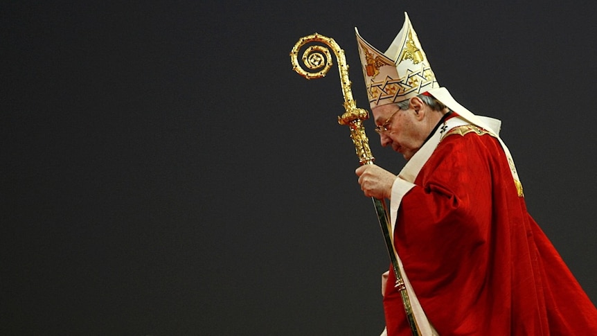 Pell, wearing red religious robes, looks down as he carries an ornate staff.