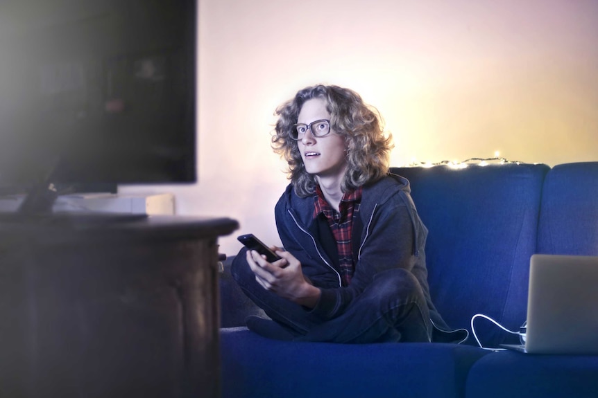 A young man with long blond hair and wearing glasses is watching a TV, leaning forward like he's captivated by the show.