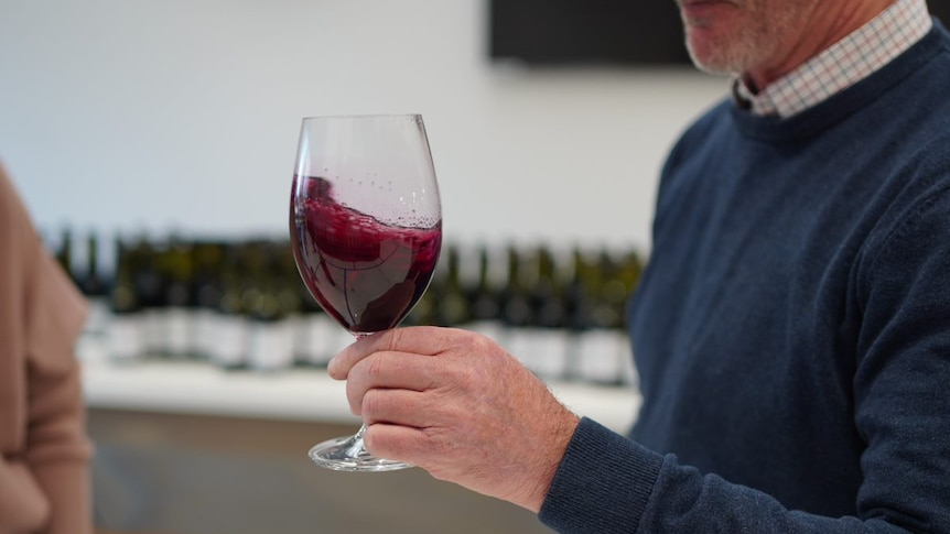 A close up of a man's hand swirling a glass of red wine in front of a row of wine bottles