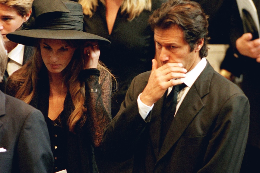 Jemima in a black dress and hat is with her then husband, Imran, who is wearing a black suit. He has his hand over his mouth.