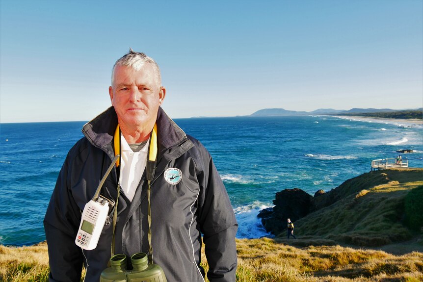 older man with walkie talkie and binoculars facing camera with ocean in background