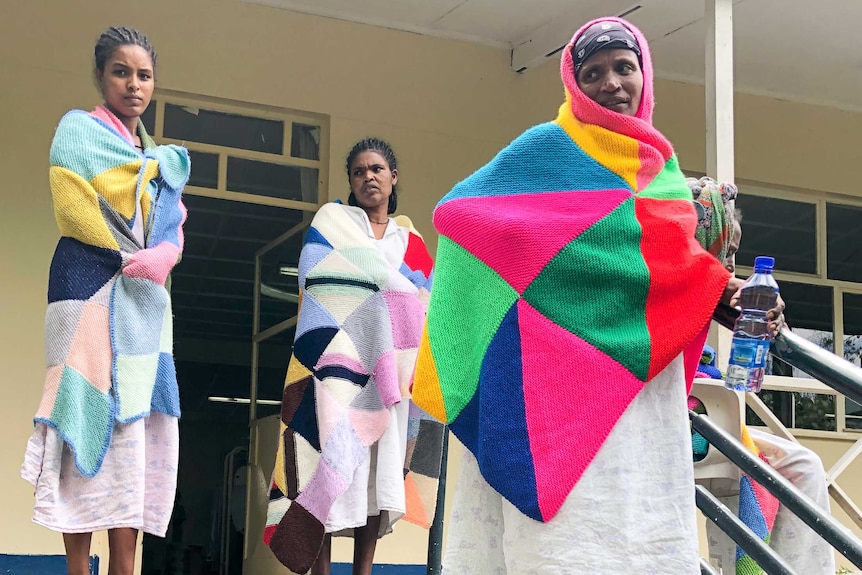 Four women grouped together on a set of stairs, wrapped in brightly-coloured knitted blankets. One women has a catheter.