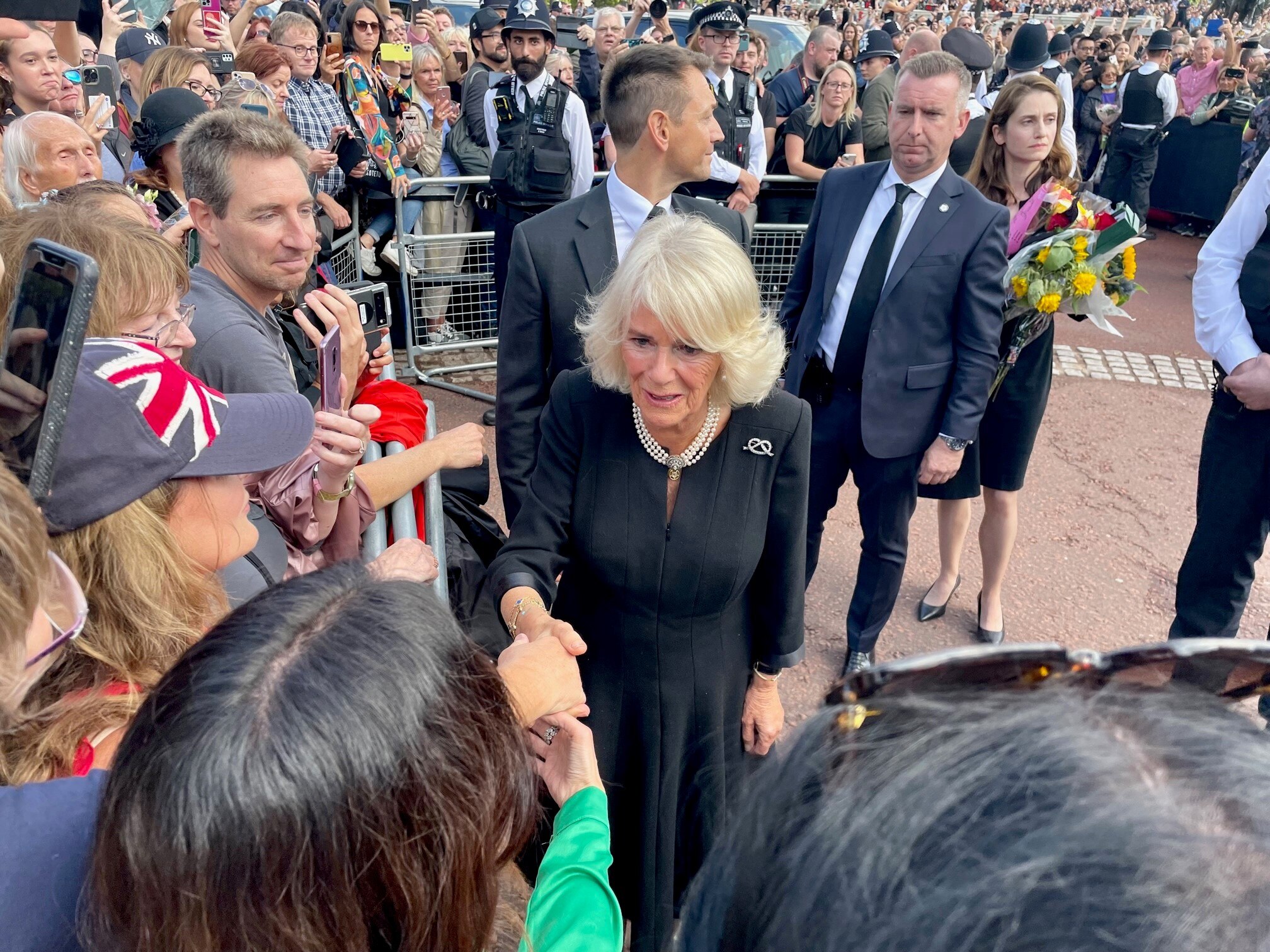 Camilla in a black dress shakes hands with people in a crowd 
