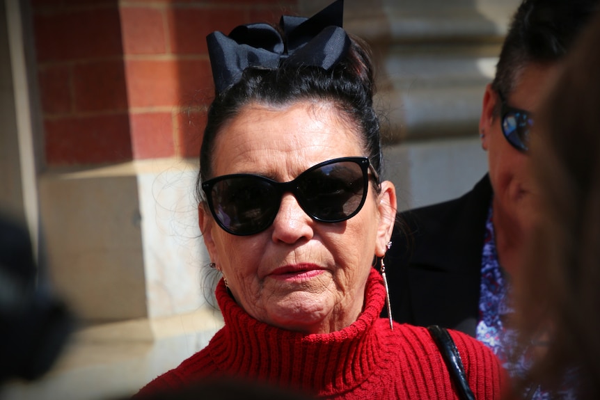 Une femme d'âge moyen portant des lunettes de soleil, un pull rouge et un nœud noir dans ses cheveux.